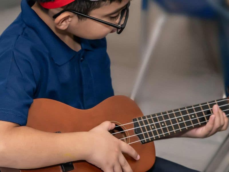 A young student in glasses strums a ukulele during a lesson